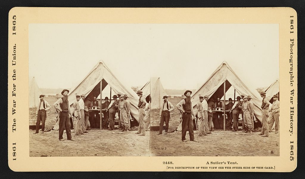 A Civil War sutler standing outside a tent while soldiers queue inside